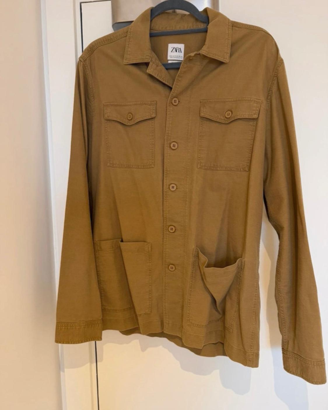 Buttoned brown jacket hanging on a hanger on a white door. It has four front pockets. ZARA can be read on the label.