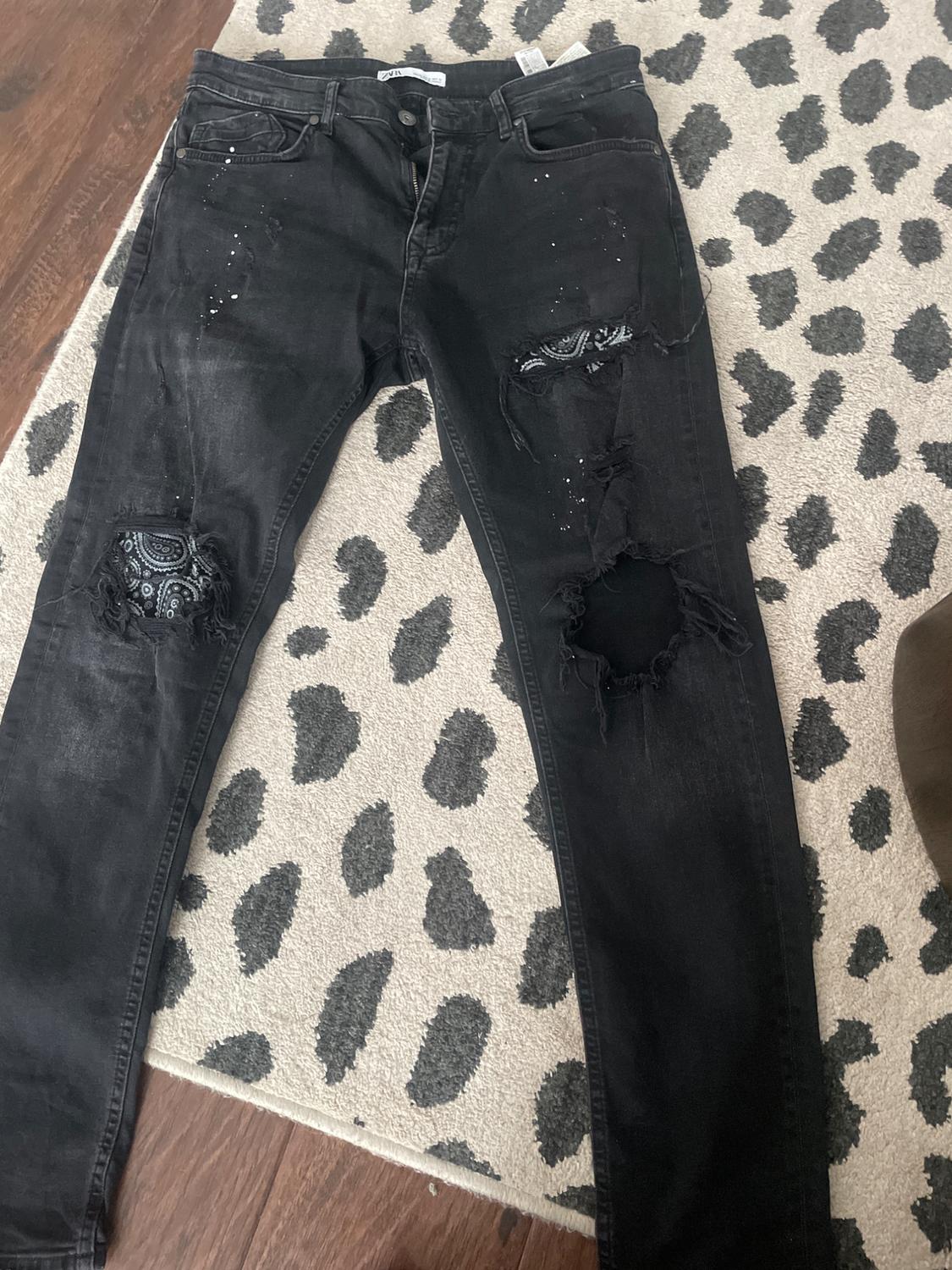 Black ripped jeans with bandana-print patches, white splatters, and ZARA label, on a carpet with black spots.