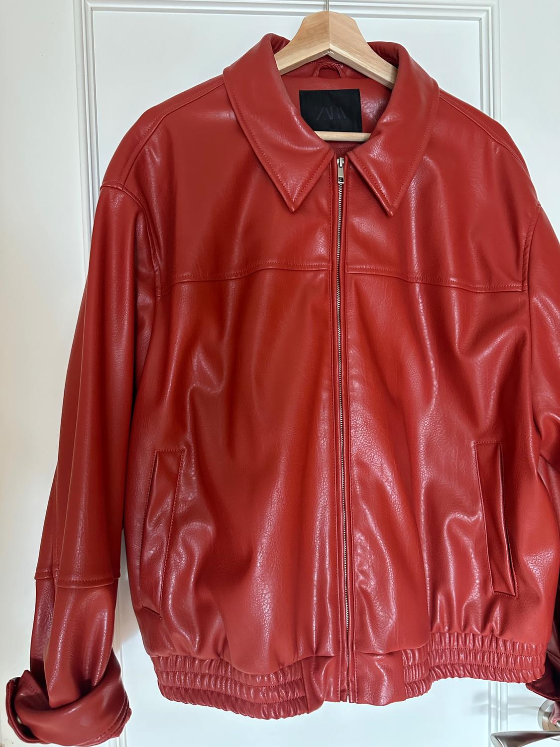 Κόκκινο jacket από συνθετικό δέρμα κρεμασμένο σε ξύλινη κρεμάστρα, με φερμουάρ μπροστά και πλαϊνές τσέπες. Λευκό φόντο.