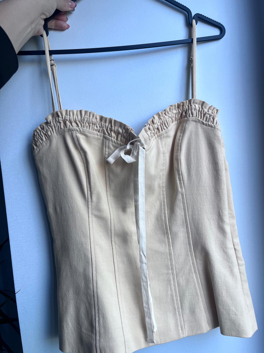 Top in beige with thin straps and ruched detailing on the neckline, hanging on a black hanger against a white background.