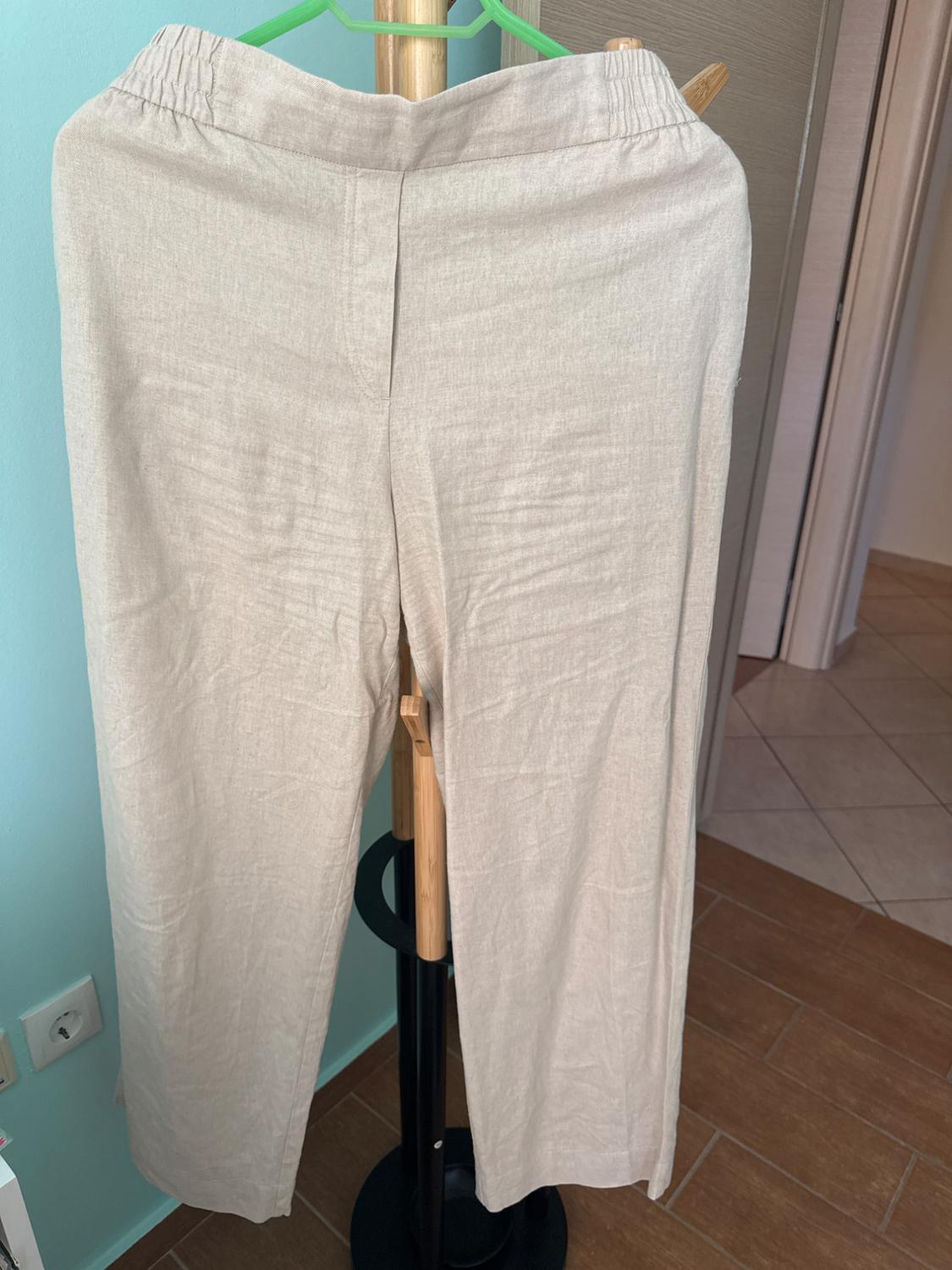 Beige linen trousers hanging on a green hanger against a blue wall background and brown tiled floor.
