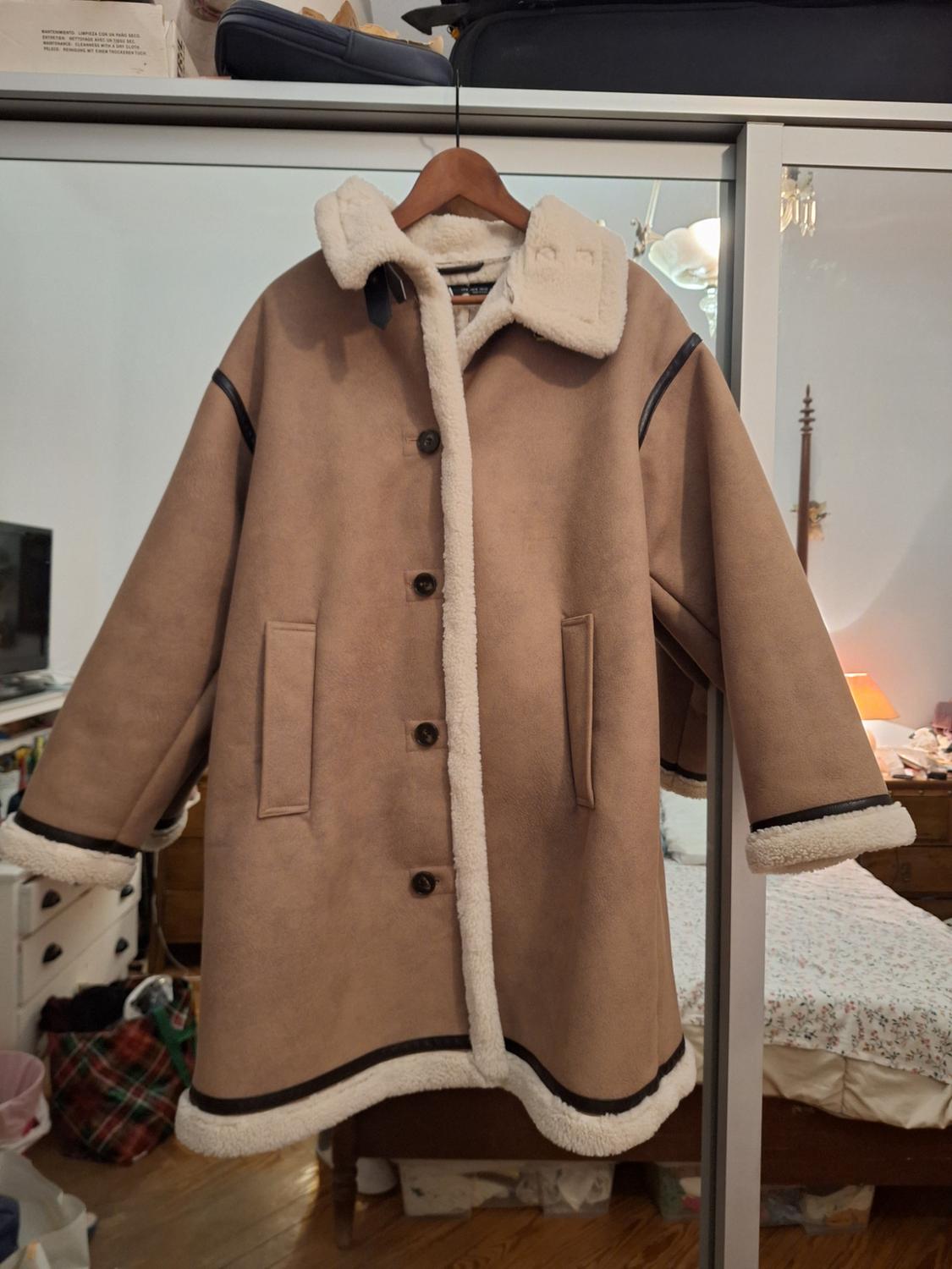 Brown coat with white lining hanging on a wooden hanger in a bedroom. Background with bed and lit lamp.