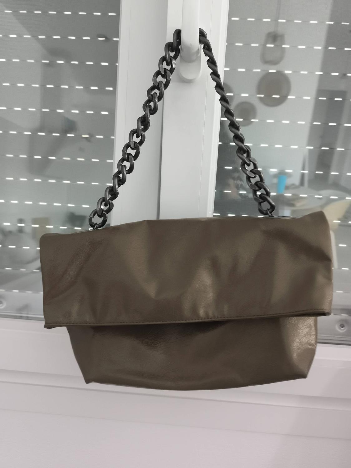 Brown handbag hanging from a black chain against a window background with white blinds. Modern and minimalist atmosphere.