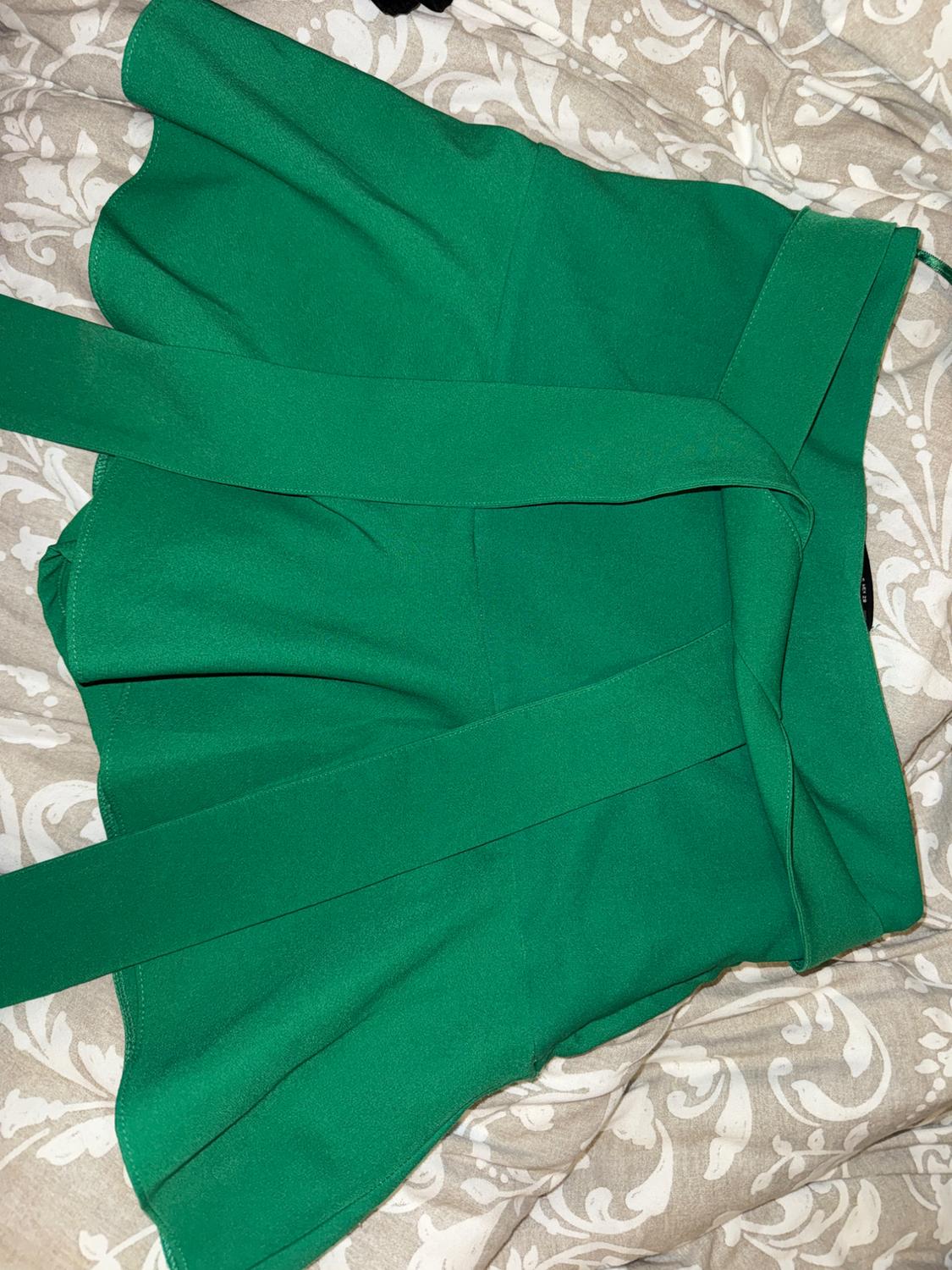 Green skirt with a wide belt on fabric with a floral design in beige and white tones. The skirt is laid out on the fabric.