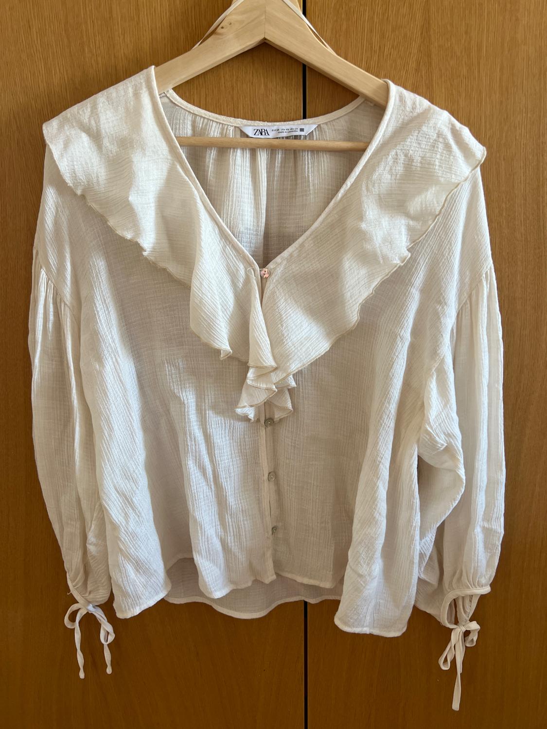 White blouse with a V-neck, frills, buttons and ribbon ties at the cuffs. ZARA text on the label. Hanging on a hanger against a wooden background.