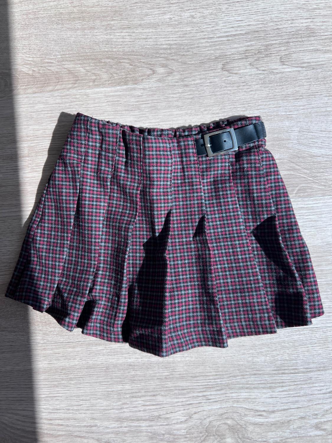 Pleated skirt with a plaid pattern in red and gray tones, with a black side stripe and buckle, on a light wood surface.