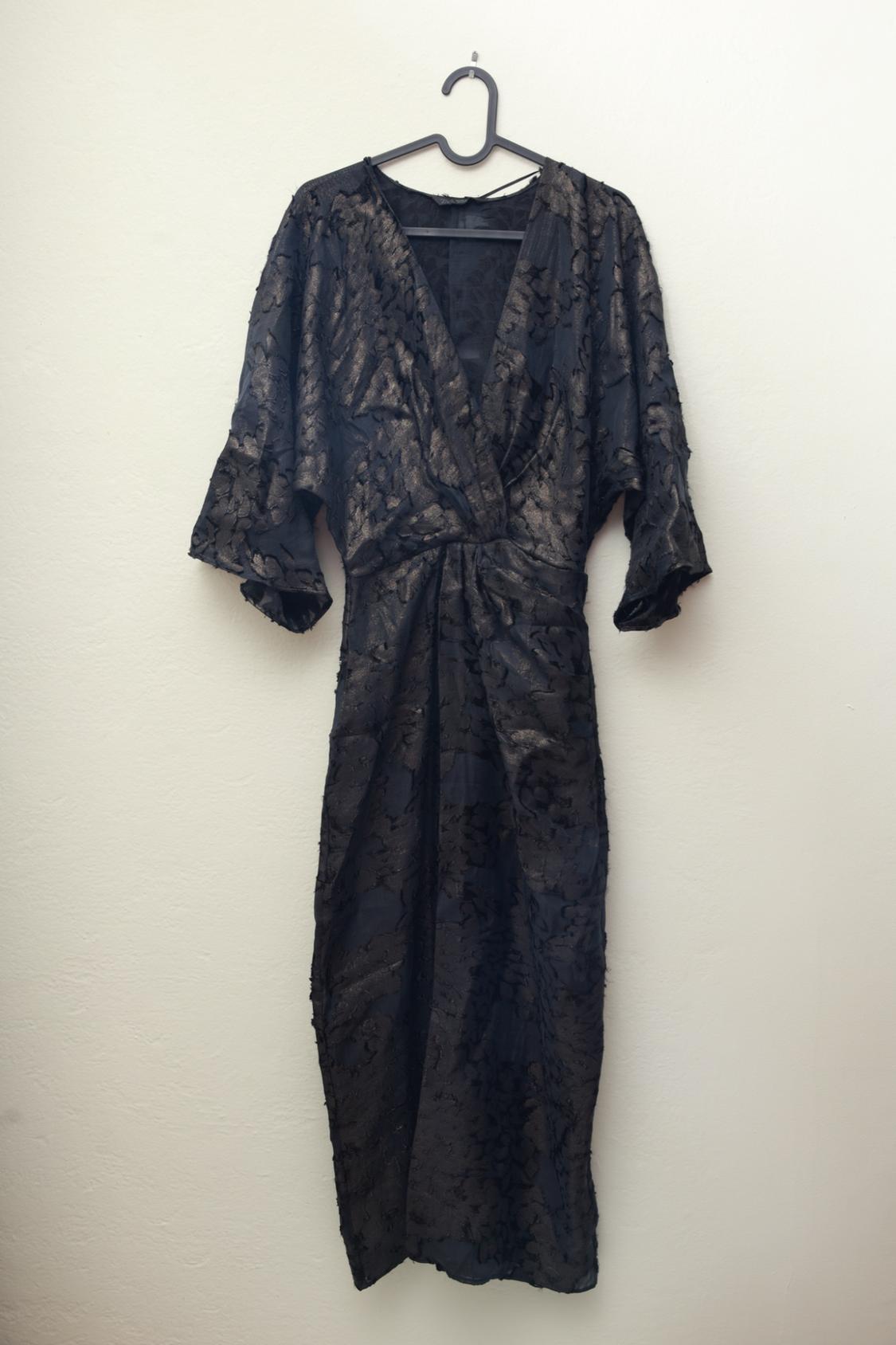Black dress with gold texture, V-neck, and wide short sleeves,
hanging on a black hanger against a white background.