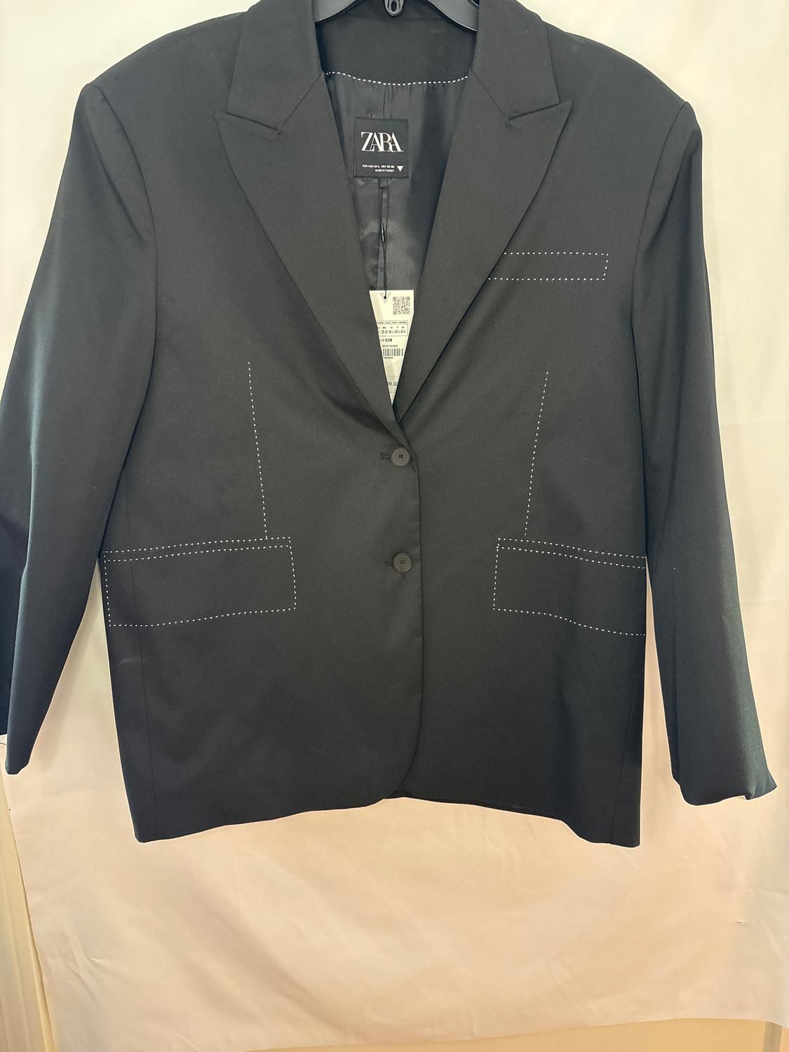 Black suit jacket hanging on a hanger, with white stitching details and a visible tag on the inside. ZARA brand.