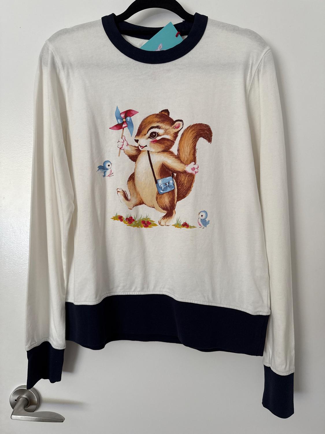 White t-shirt with dark blue trim hanging on a door. It features a drawing of a squirrel with a windmill next to two blue birds.