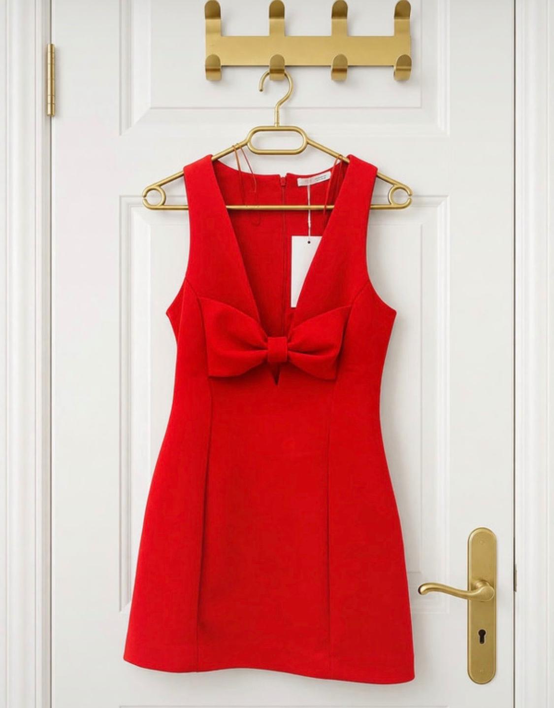 Short red sleeveless dress with a deep V-neckline and large front bow, hanging on a gold coat hanger over a white door.