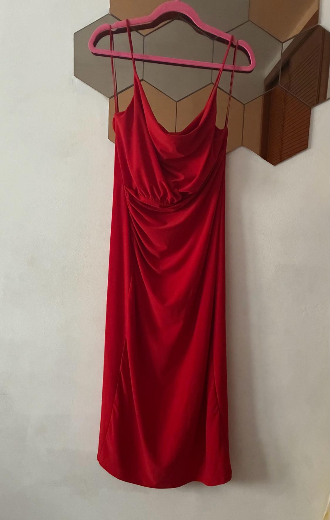 Red floor-length spaghetti strap dress with front draping, hanging on a pink hanger on a hexagonal mirror against a white wall.