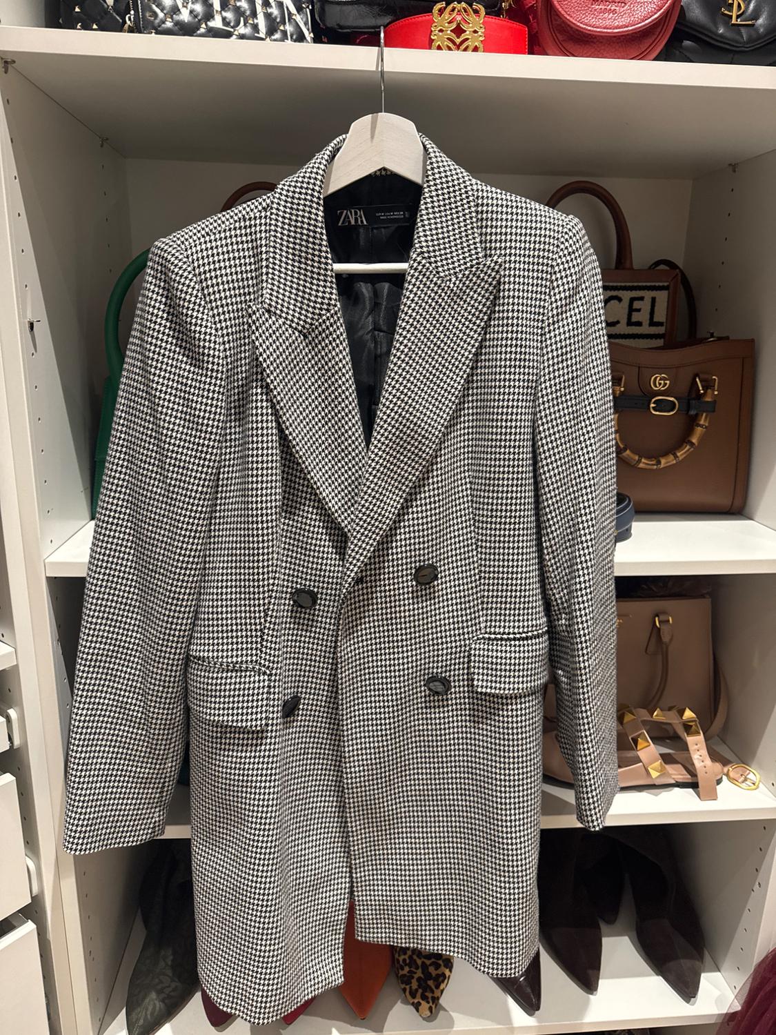 Black and white checked coat hanging in a wardrobe, surrounded by luxury handbags on shelves. Visible label: ZARA.