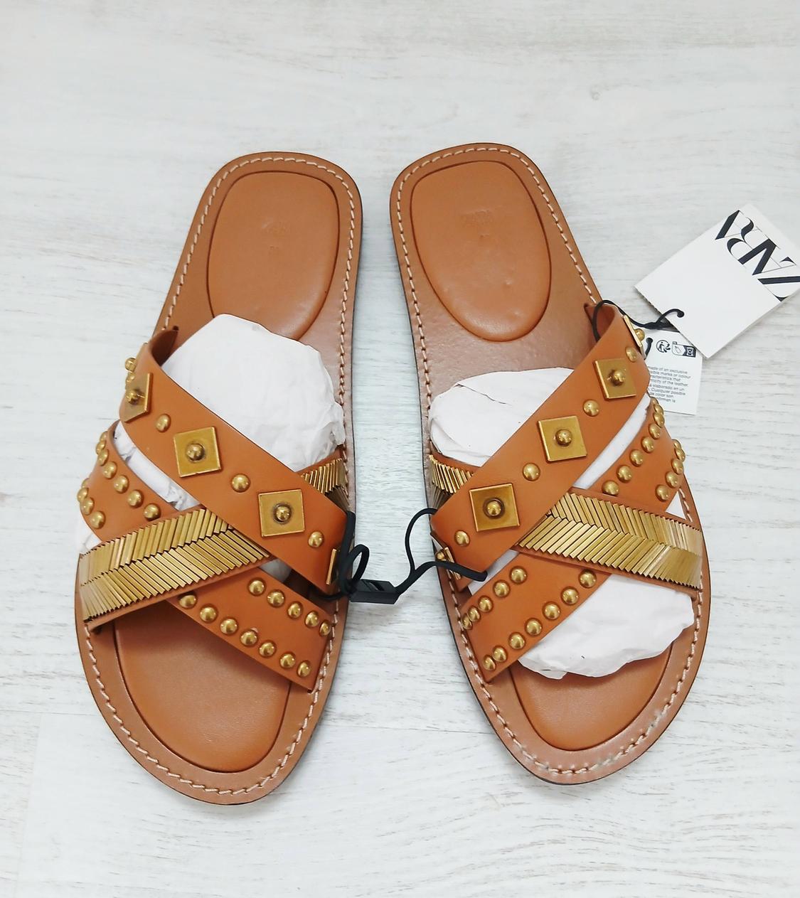 Brown sandals with crossed straps decorated with gold studs and embossed details. Visible label with "ZARA" text.