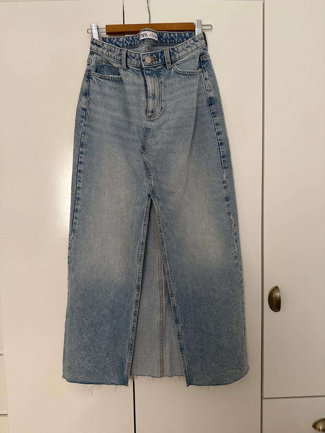 Light blue denim skirt with a front split, hanging on a wooden hanger in front of a white wardrobe. Visible label: "ZARA".