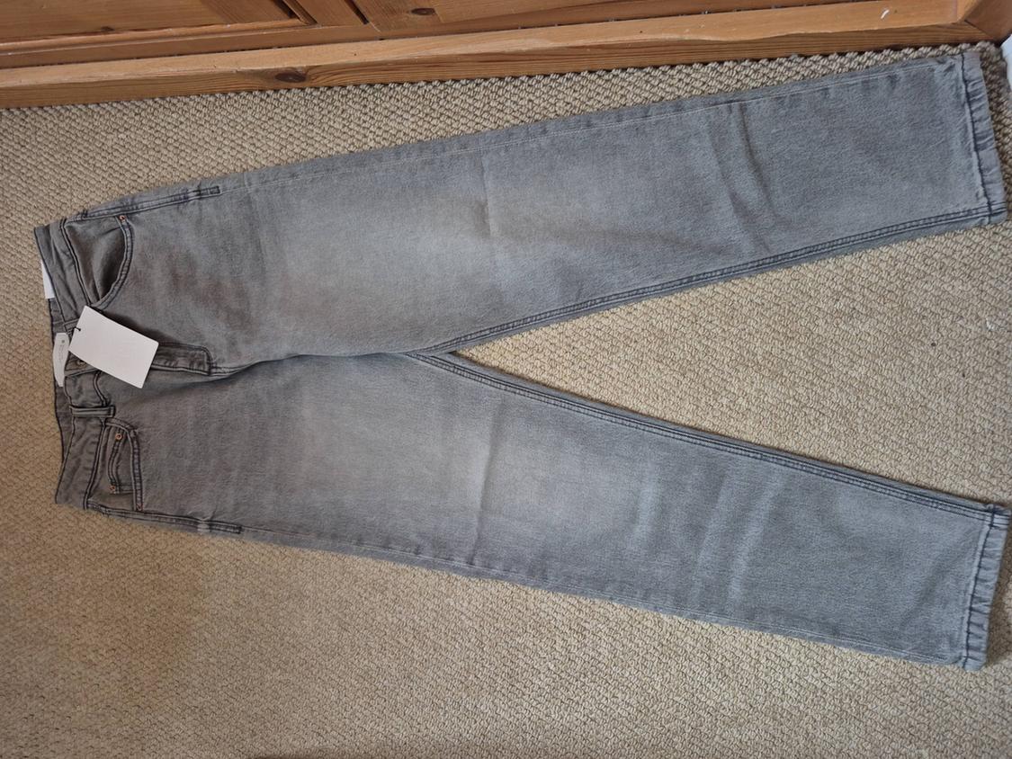 Grey denim jeans spread out on a beige rug, with a white tag on the left back pocket.