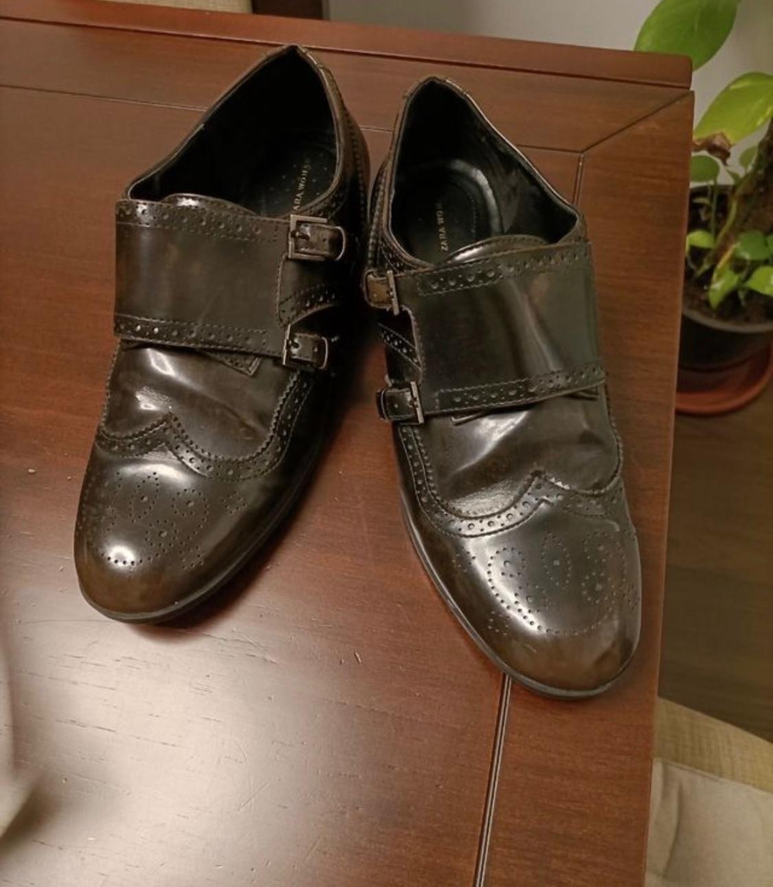Brown leather double monk strap shoes with punched detailing on a wooden table next to a plant. Text: ZARA WOMAN.
