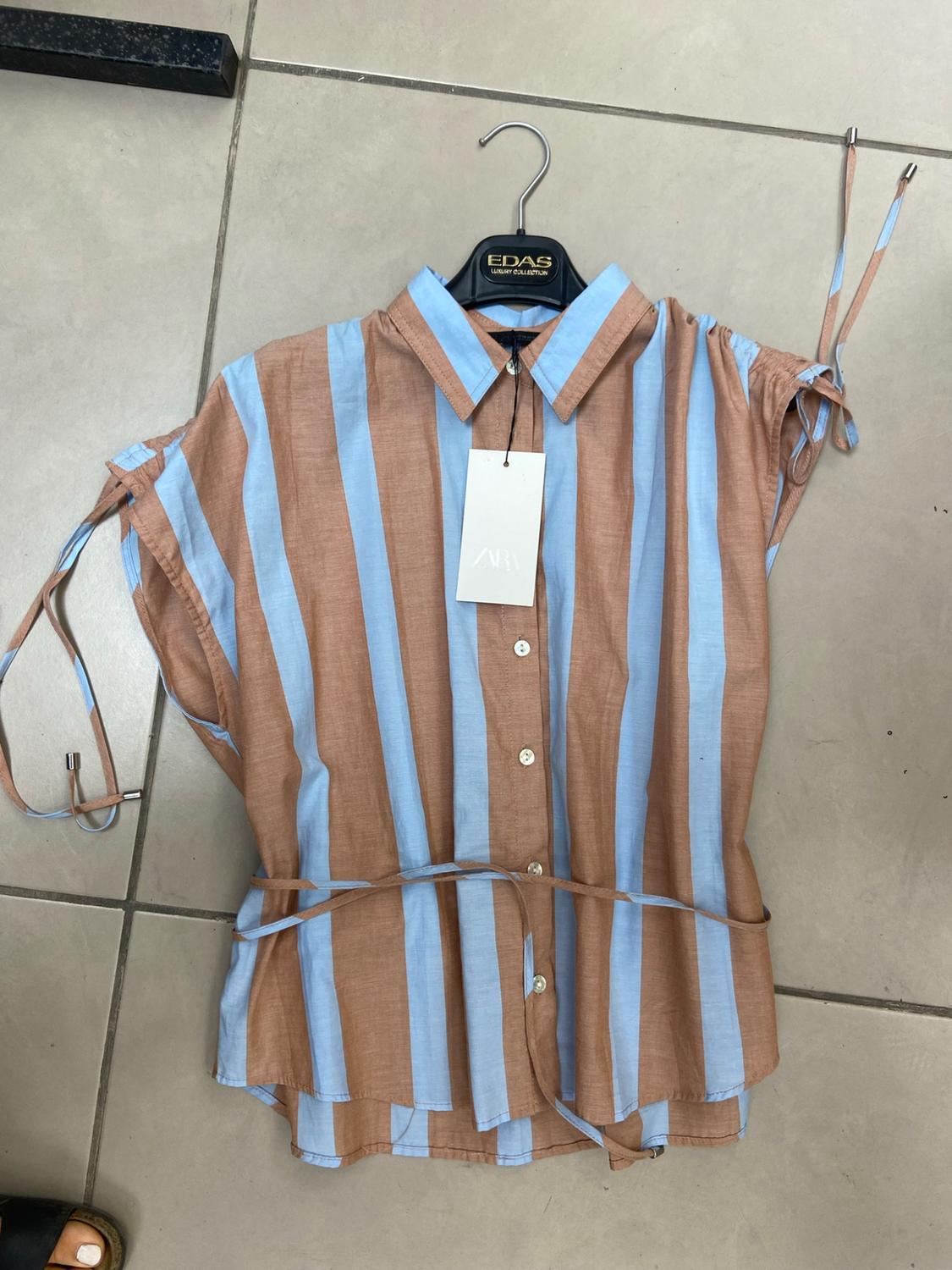 Blue and brown striped shirt with buttons. Hanger with the text EDAS LUXURY COLLECTION and a ZARA label on a tiled floor.