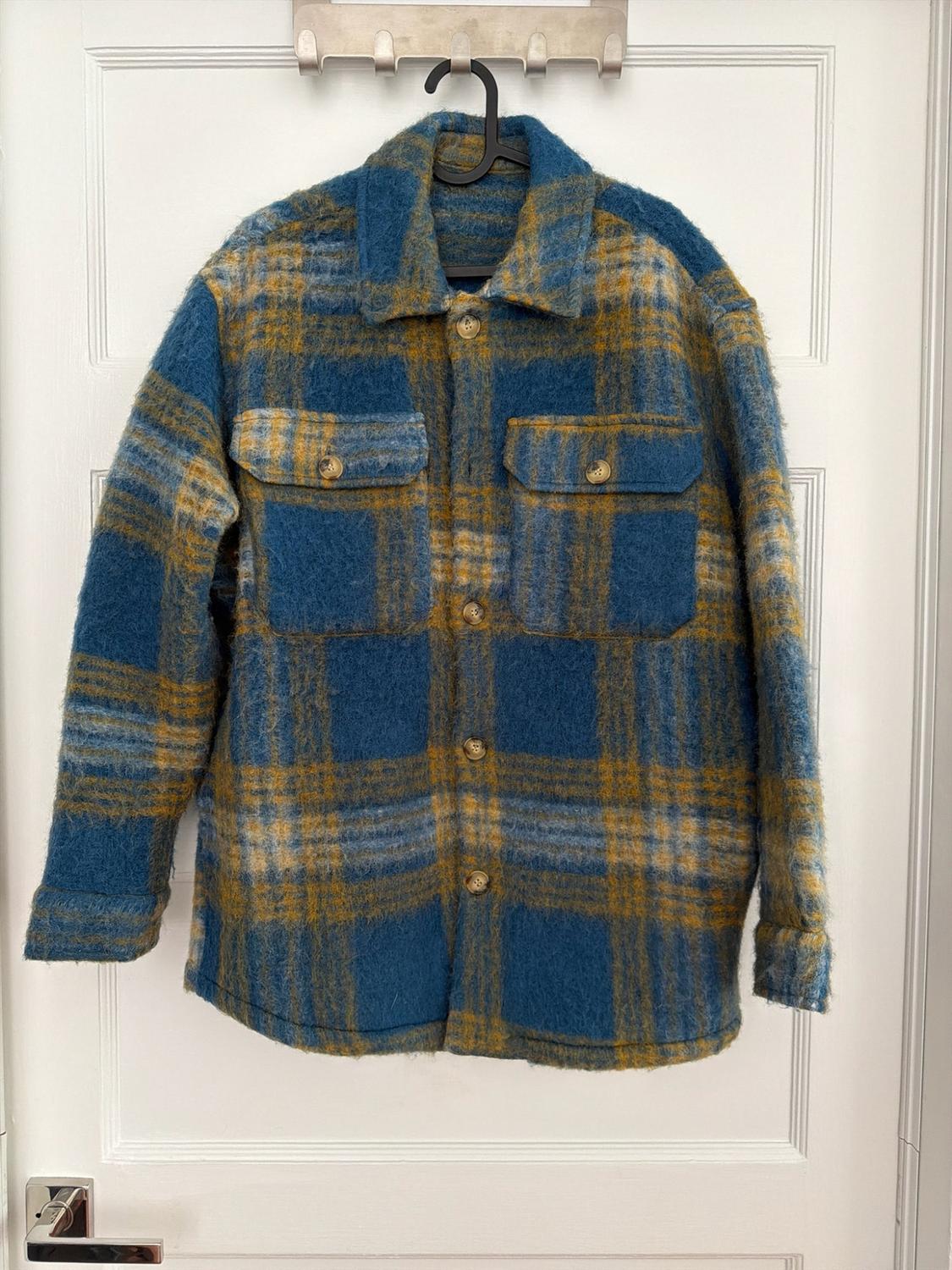 Blue and yellow plaid overshirt with front pockets, hanging on a hanger over a white door.