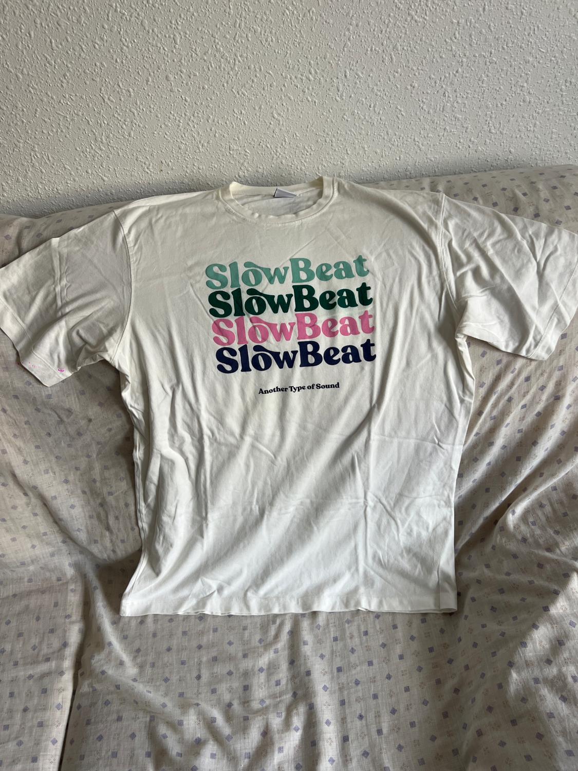 White t-shirt on a bed, with "SlowBeat" text repeated in colours and "Another Type of Sound" in black. White wall background.