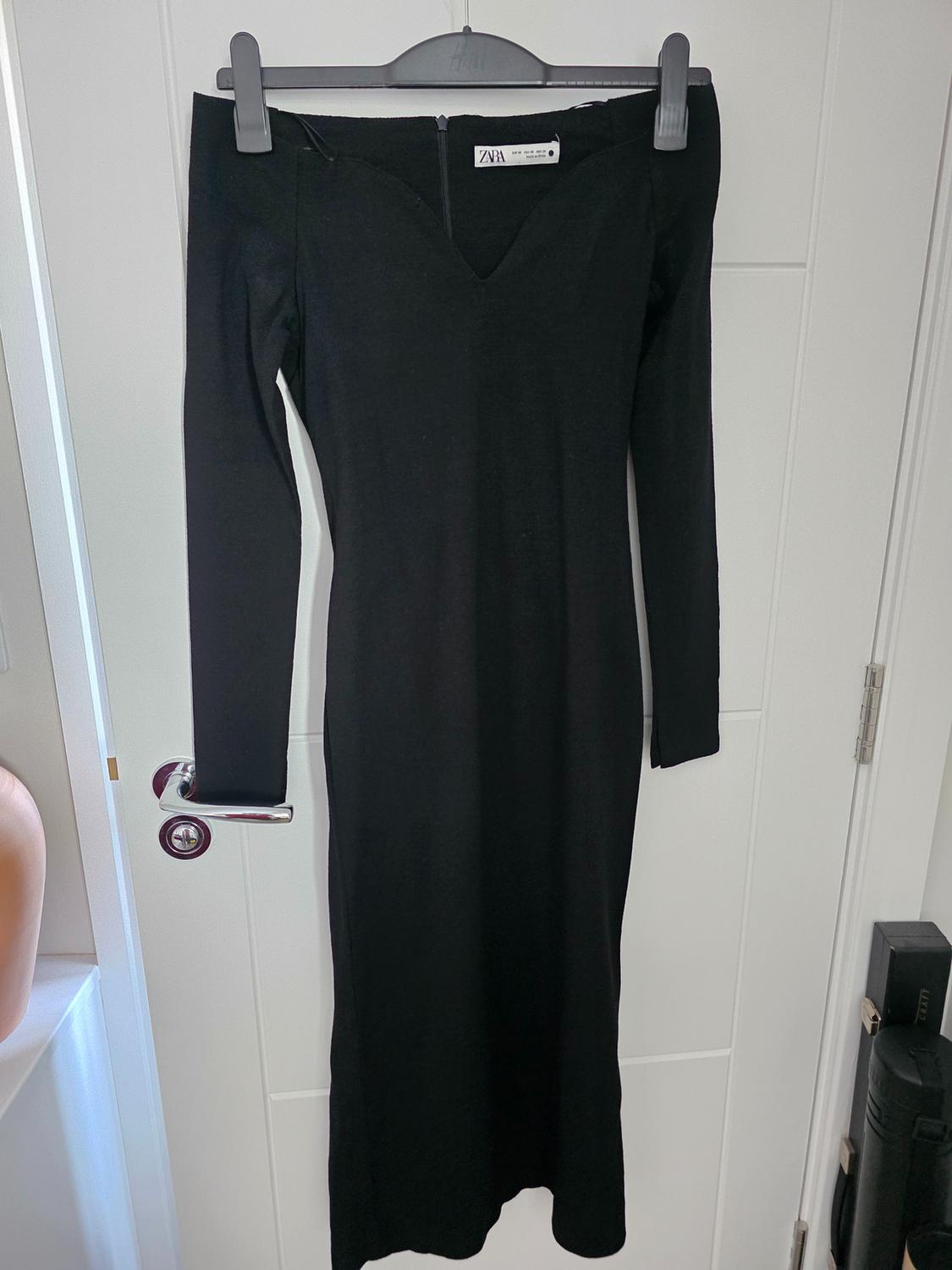 Black long-sleeved dress hanging on a grey hanger. Visible label with the text "ZARA". White door background.