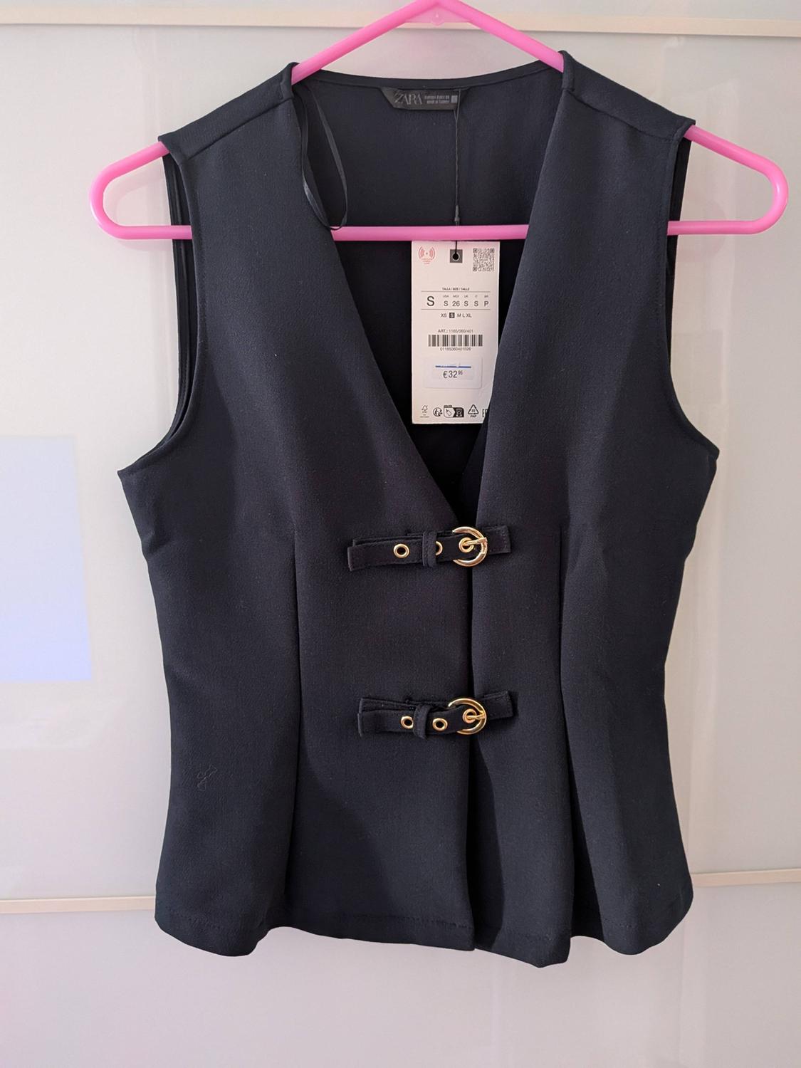 Black sleeveless waistcoat with two gold buckles, hanging on a pink hanger. Visible tag with price of €32.