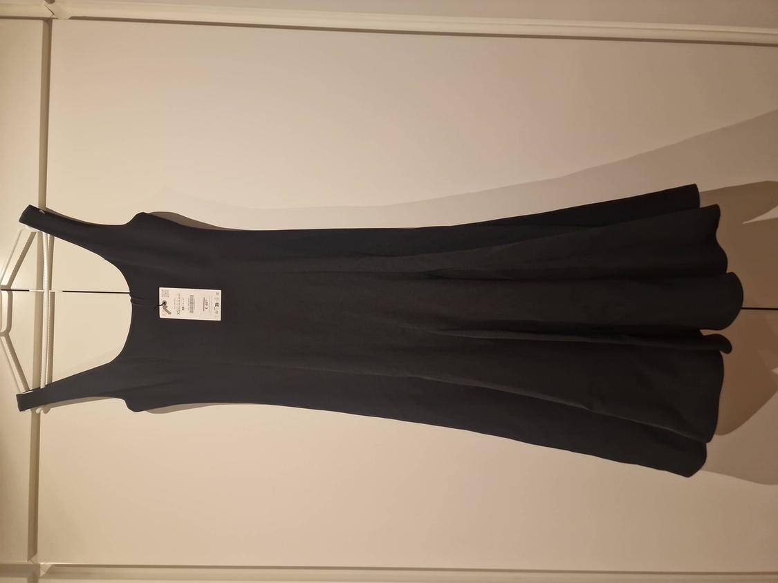 Black, sleeveless dress with a flared skirt, hanging on a hanger over a white wardrobe. A white tag is visible on the chest.