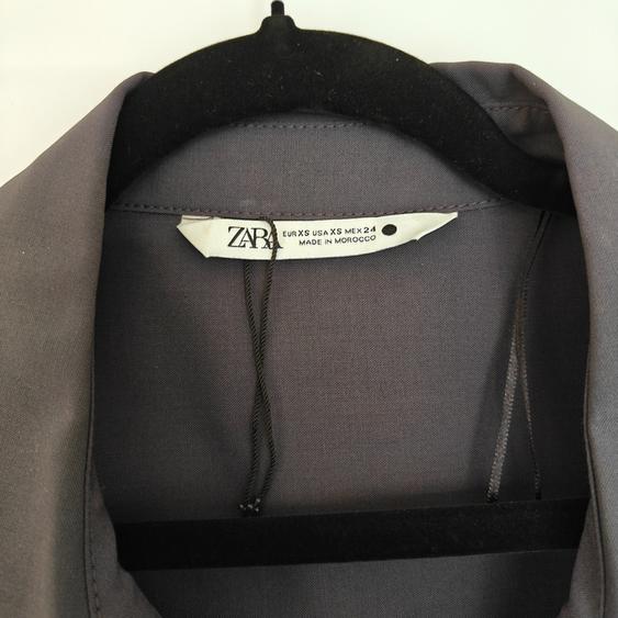 Label of a grey garment on a black hanger. Visible text: "ZARA, EUR XS USA XS MEX 24, MADE IN MOROCCO".