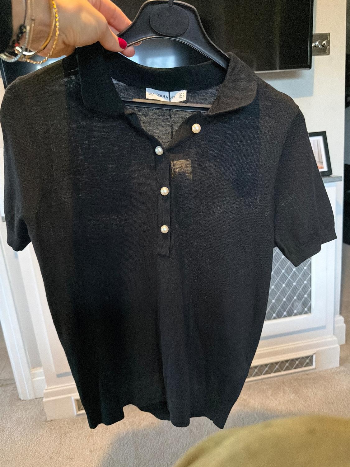 Sheer black polo shirt with pearl buttons, hanging on a clothes hanger. Interior background of a room.