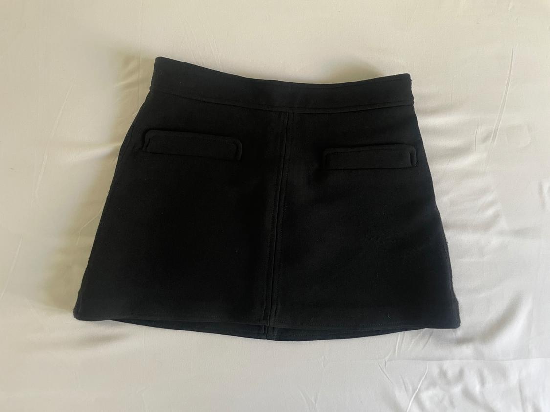 Black mini skirt with two front pockets and a central seam, photographed from above against a white fabric background.