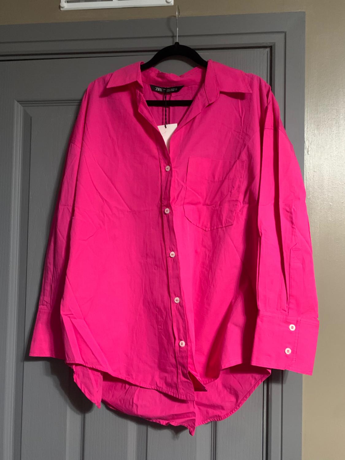 Fuchsia long-sleeved shirt hanging on a black hanger over a grey door. It has a chest pocket and white buttons.