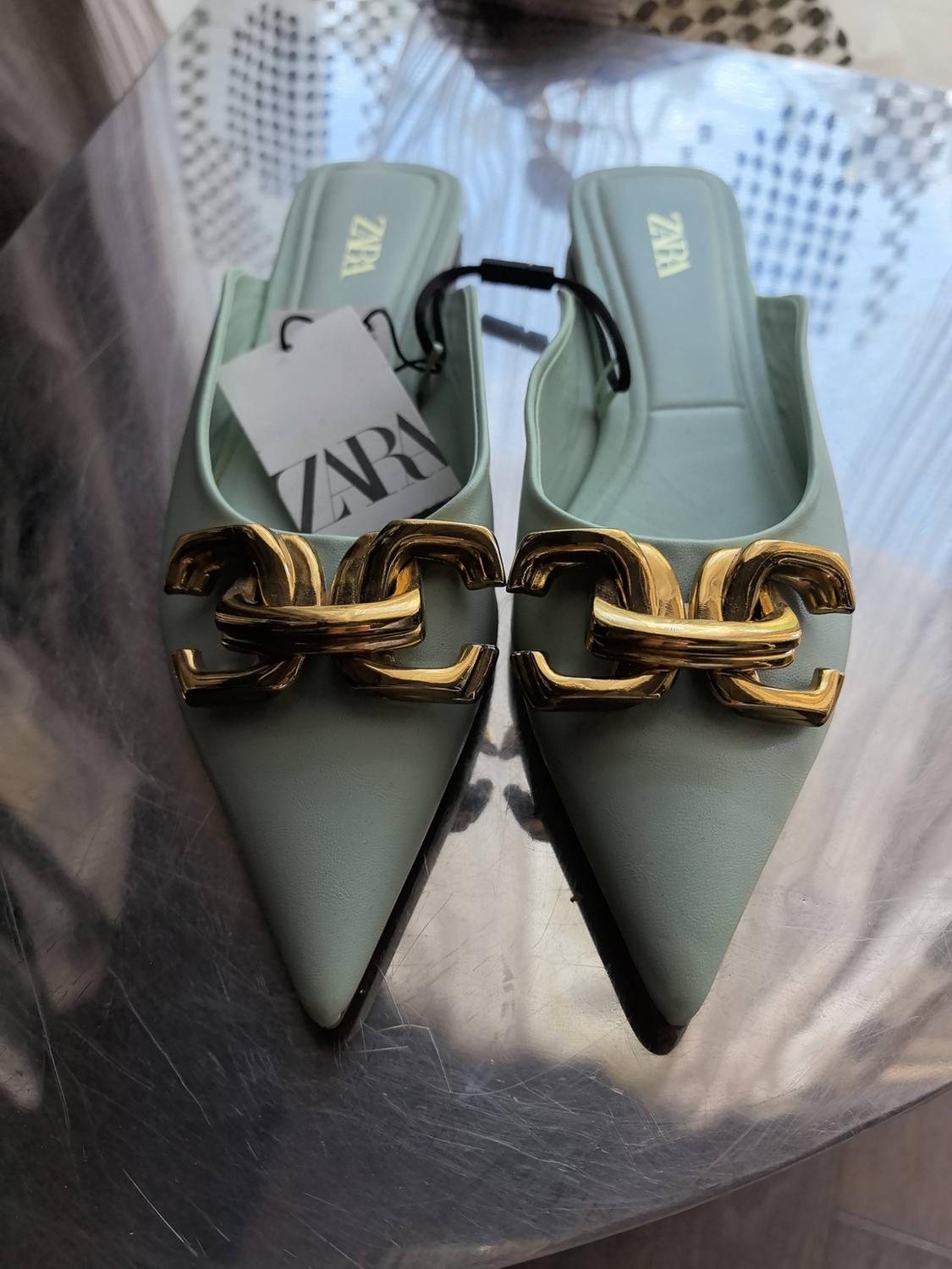 Ανοιχτοπράσινες μυτερές γόβες slingback με χρυσή αλυσίδα. Ετικέτα και εσωτερικός πάτος με κείμενο ZARA. Πάνω σε μεταλλική επιφάνεια.