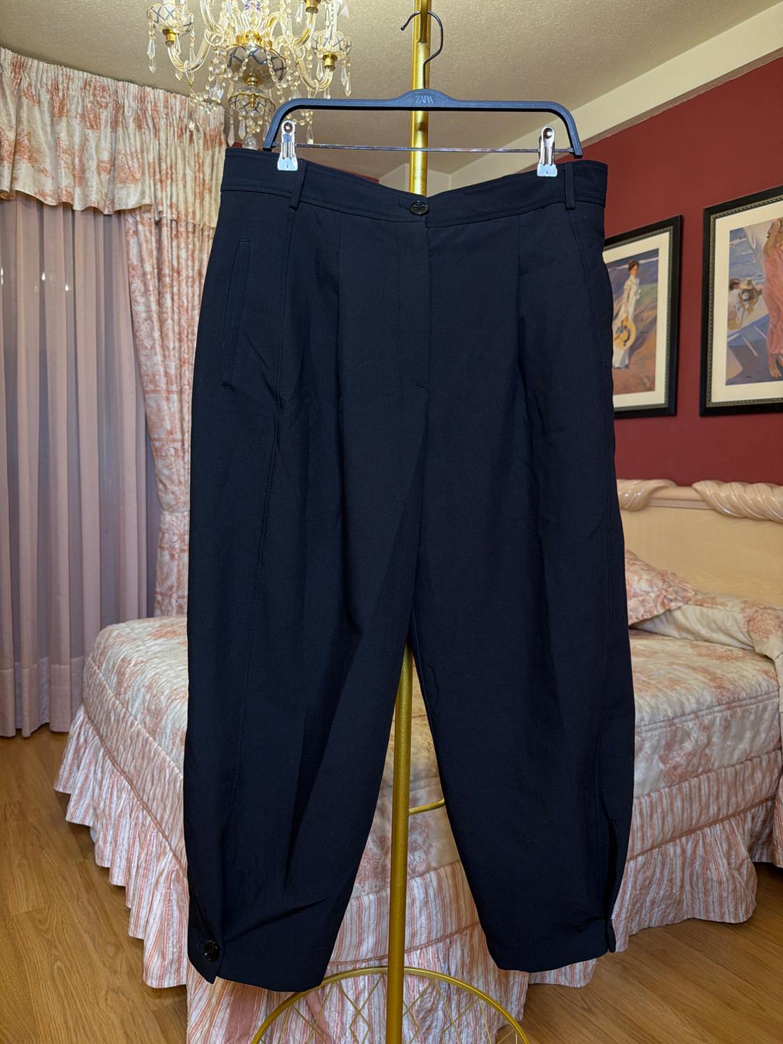 Black trousers hanging on a hanger in a bedroom with curtains and a bed with a patterned bedspread. Chandelier on the ceiling.