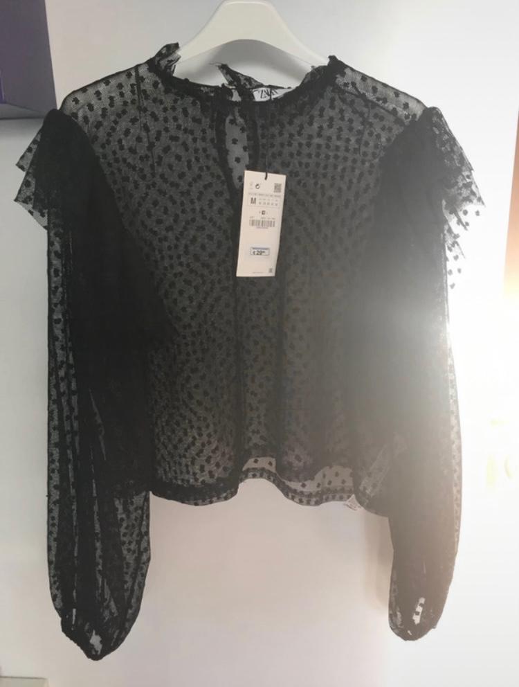 Sheer black blouse with polka dots and long puff sleeves, hanging on a white hanger. Label visible in the centre.