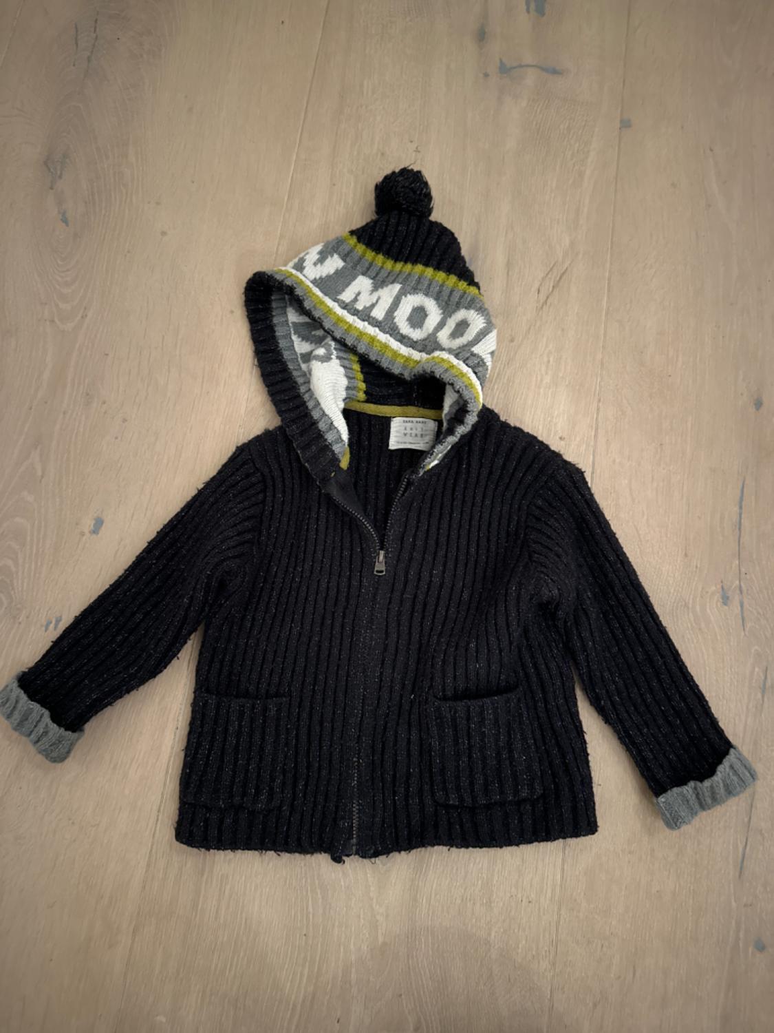 Black knitted jacket with hood and pompom, grey and green details. "MOON" text visible on the hood. Light wood background.