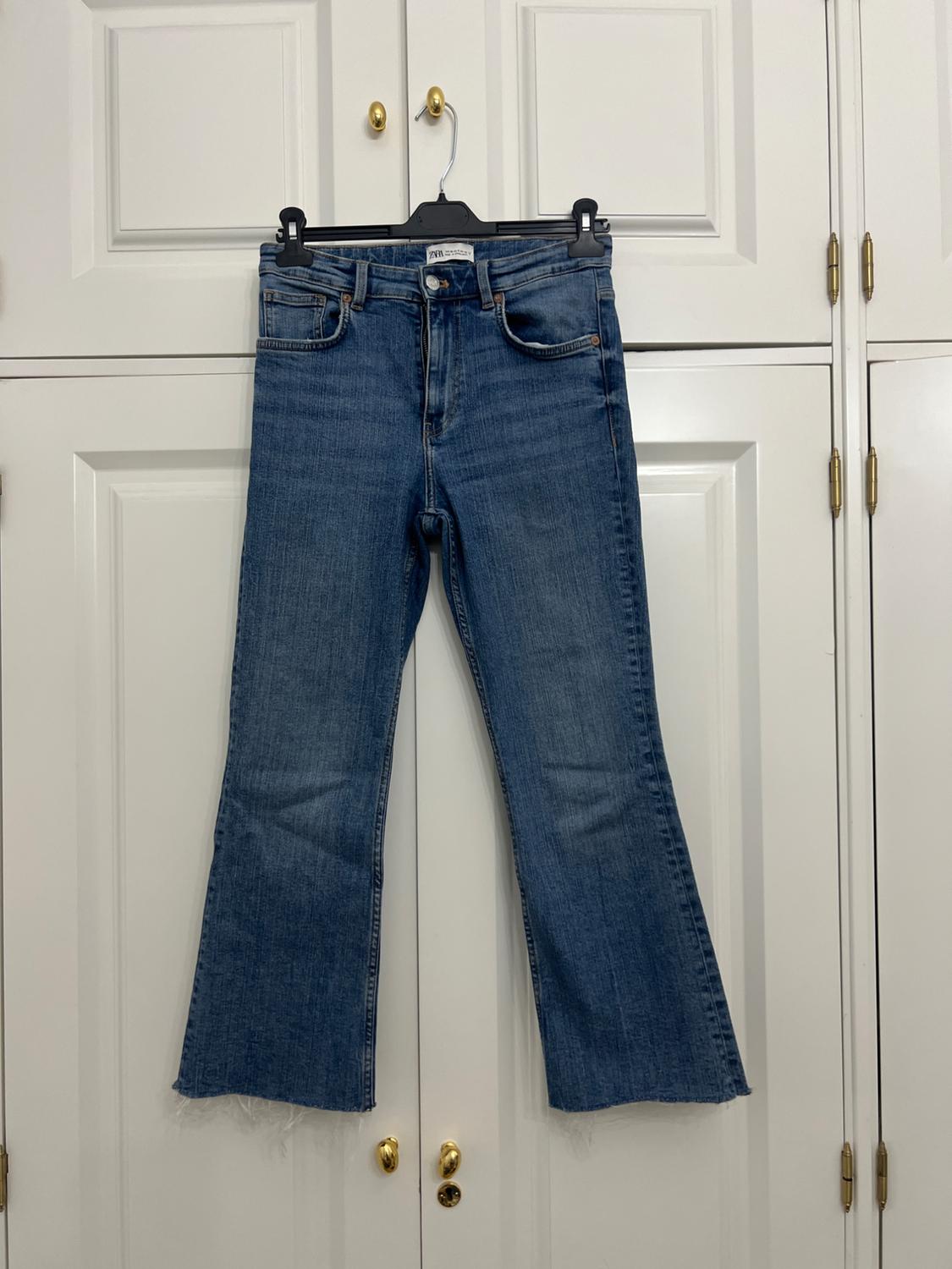 Blue denim jeans with a frayed hem, hanging on a black hanger in front of a white wardrobe with gold handles. Text: ZARA.
