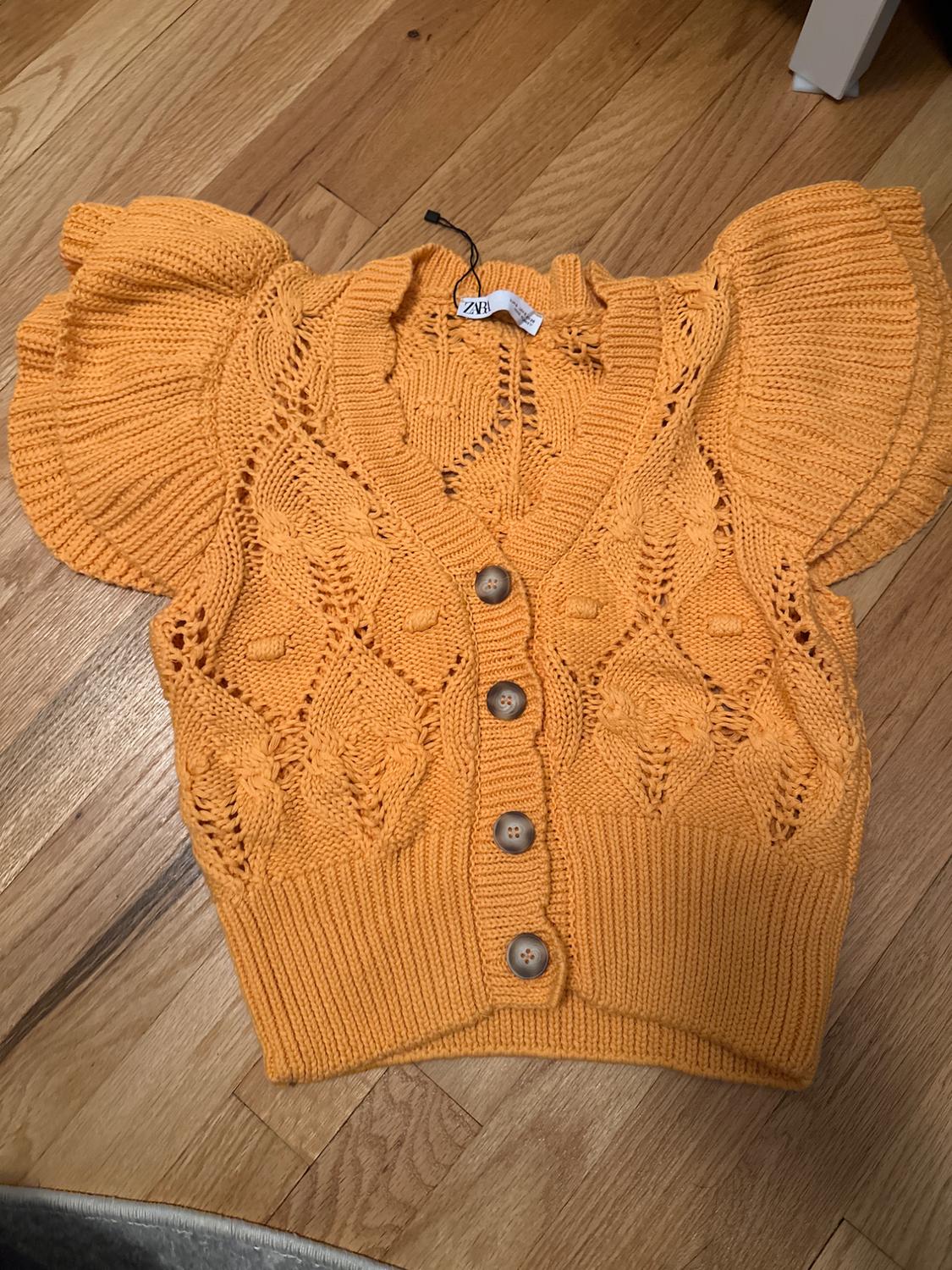 Orange short cardigan with puffed sleeves and wooden buttons on a wooden floor. Visible label with text "ZARA".