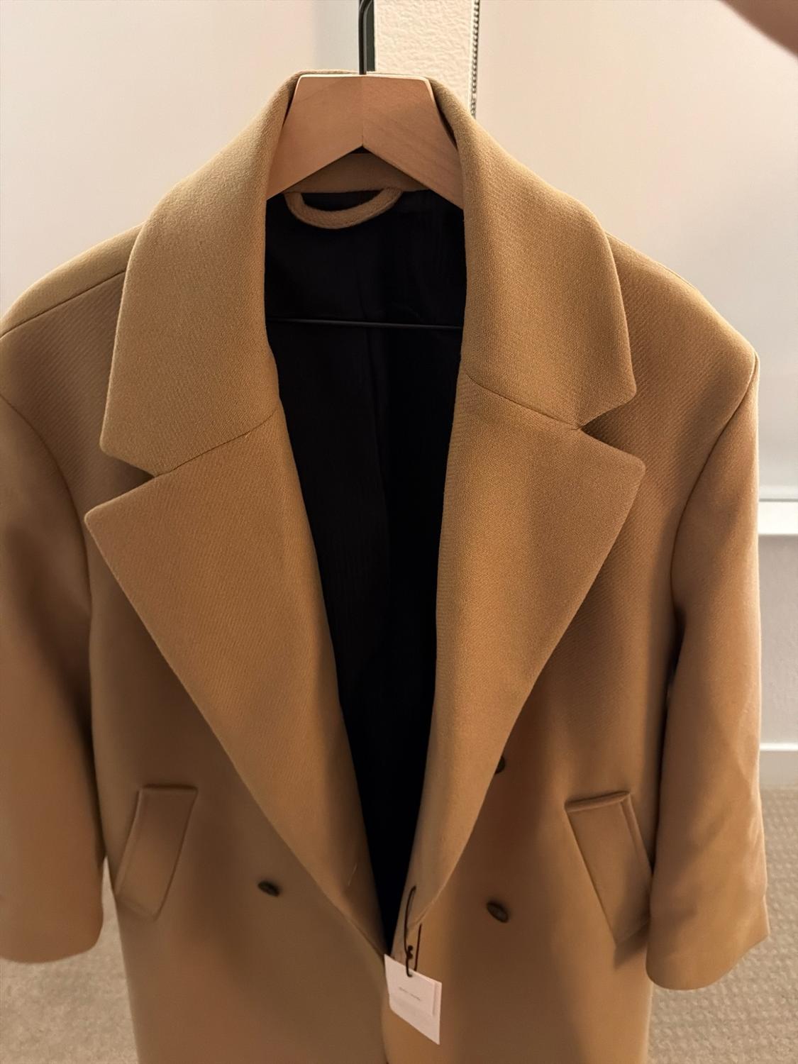 Camel wide-lapel coat hanging on a wooden hanger. It features dark buttons, a black lining, and a hanging white tag.