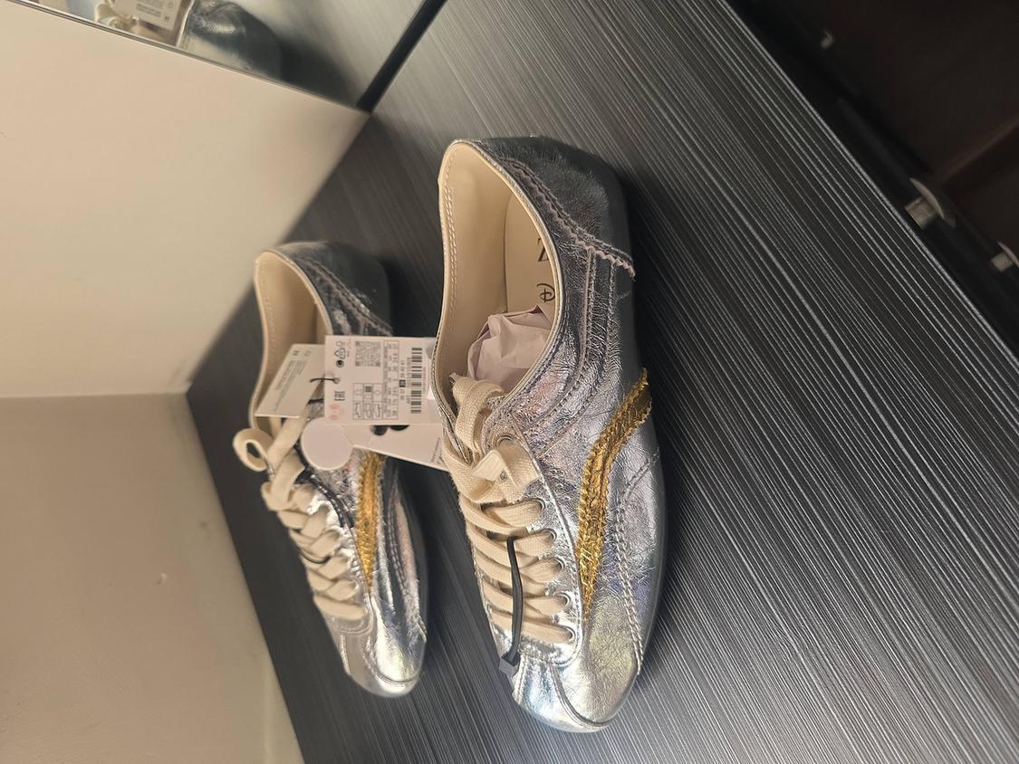 Silver trainers with gold details and a tag, on a dark surface with a mirror reflection.