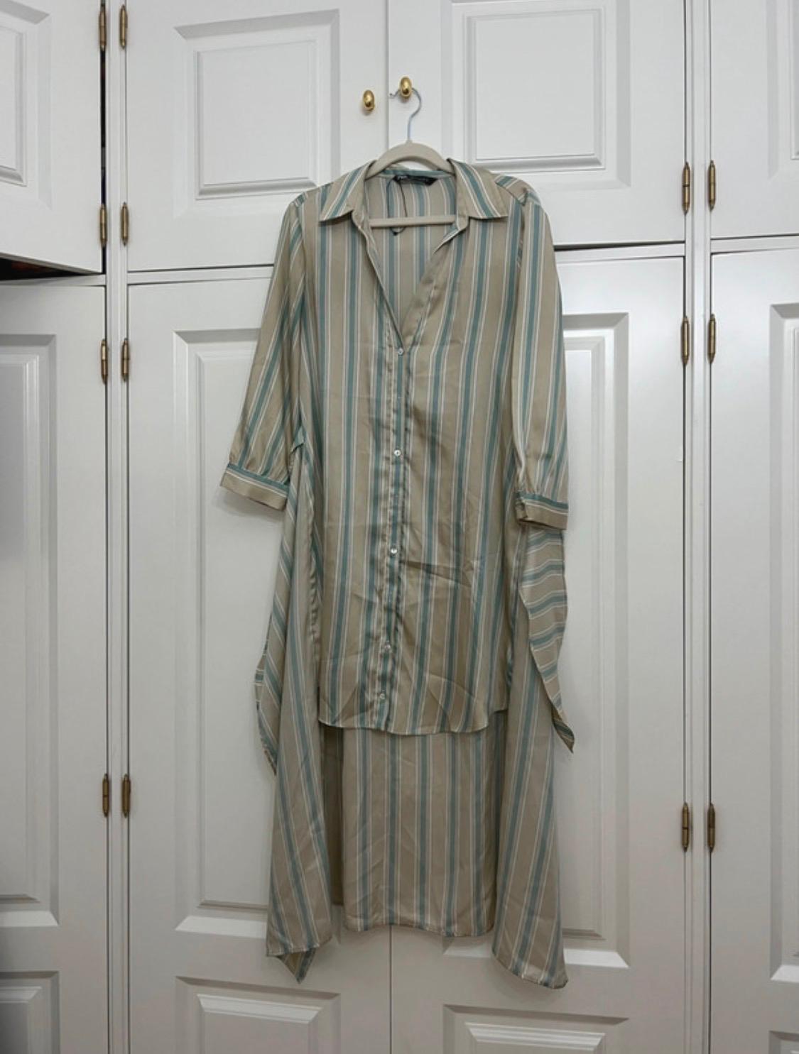 Beige and blue striped shirt dress with an asymmetric hem, hanging on the door of a white wardrobe with gold handles.