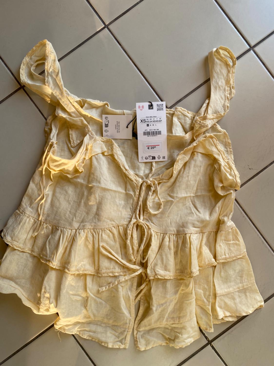 Light yellow frilled top with front ties on a tiled floor. Label with text: XS XS 24 XS XS EP €25.95.
