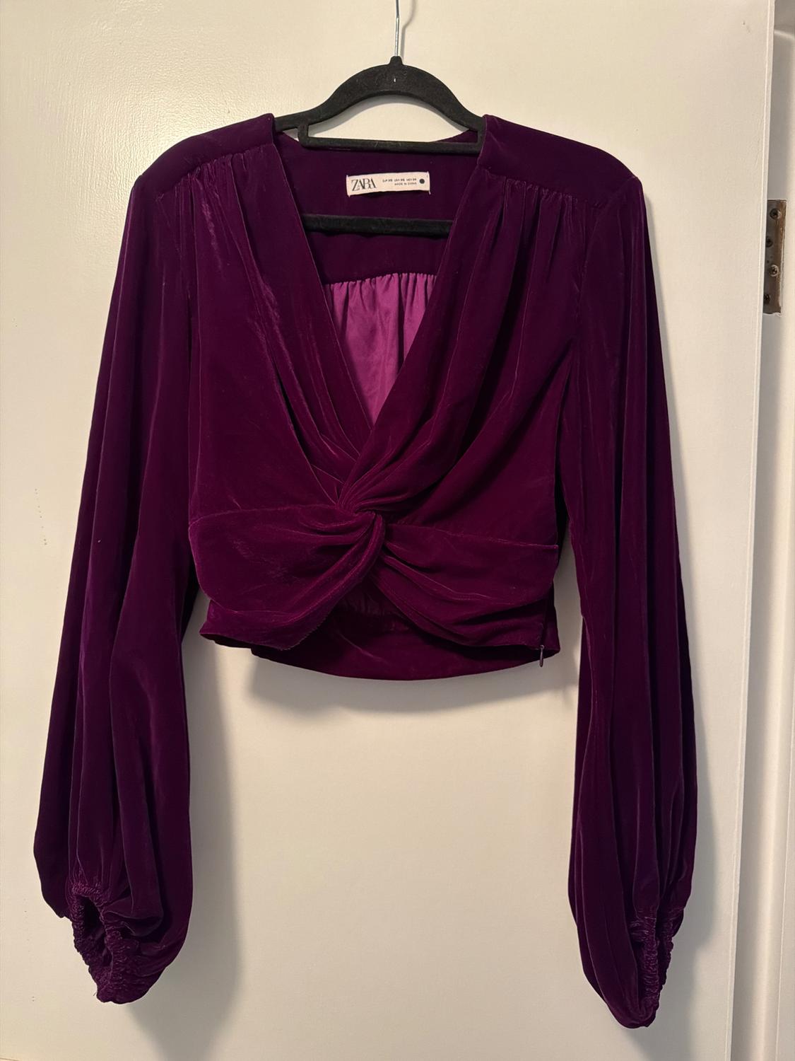 Purple velvet blouse with long puff sleeves and a wrap-over neckline, hanging on a black hanger against a white background.