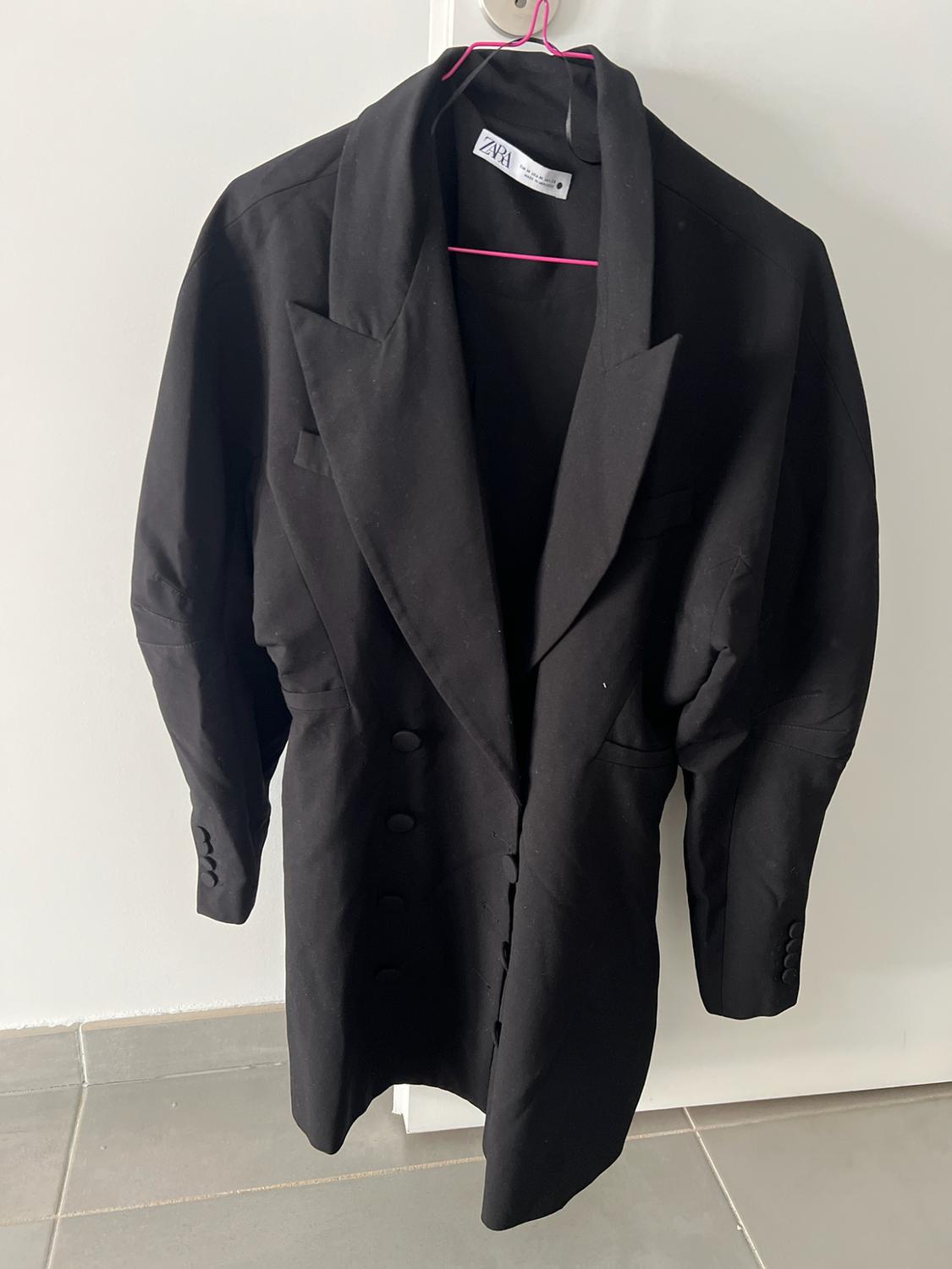 Black double-breasted blazer with structured sleeves hanging on a pink hanger over a white door. Label with the text ZARA.