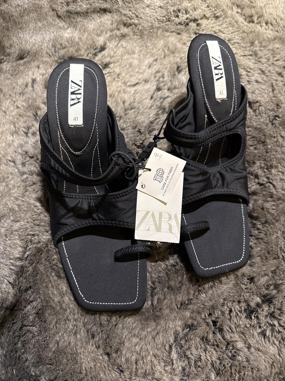 Black low-heeled sandals with a Zara tag, size 41, on a gray furry surface.