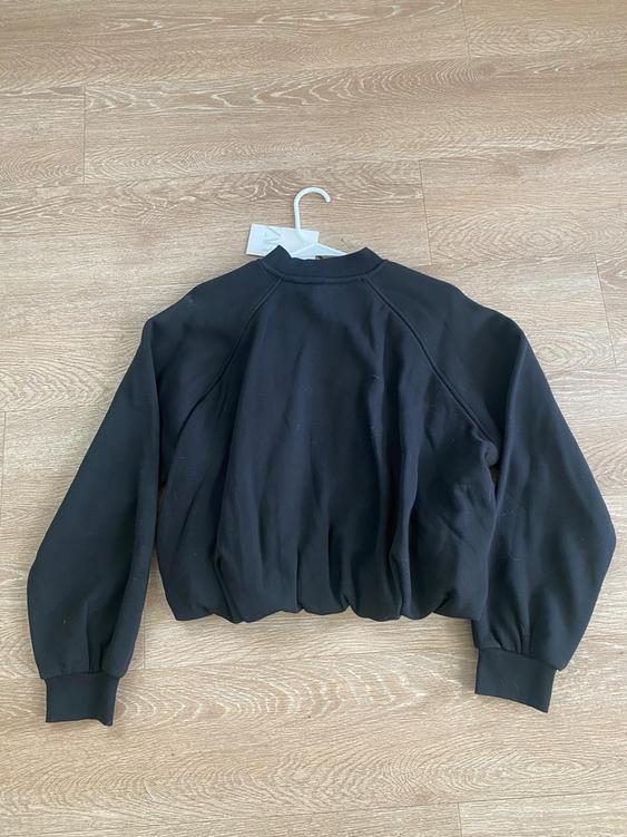 Black long-sleeved jacket with elastic cuffs, seen from behind, hanging on a hanger over a light wooden floor.