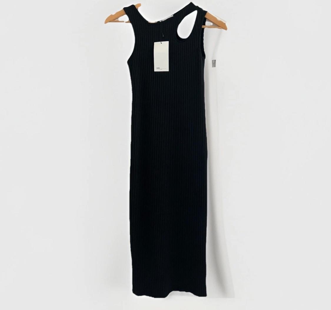 Sleeveless black ribbed maxi dress with an asymmetrical neckline and slit, hanging on a wooden hanger with a tag against a white background.