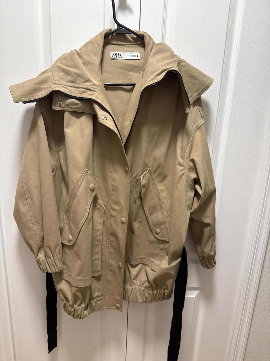 Beige jacket with a hood and large pockets, hanging on a black hanger in front of a white door. Visible tag: Zara.