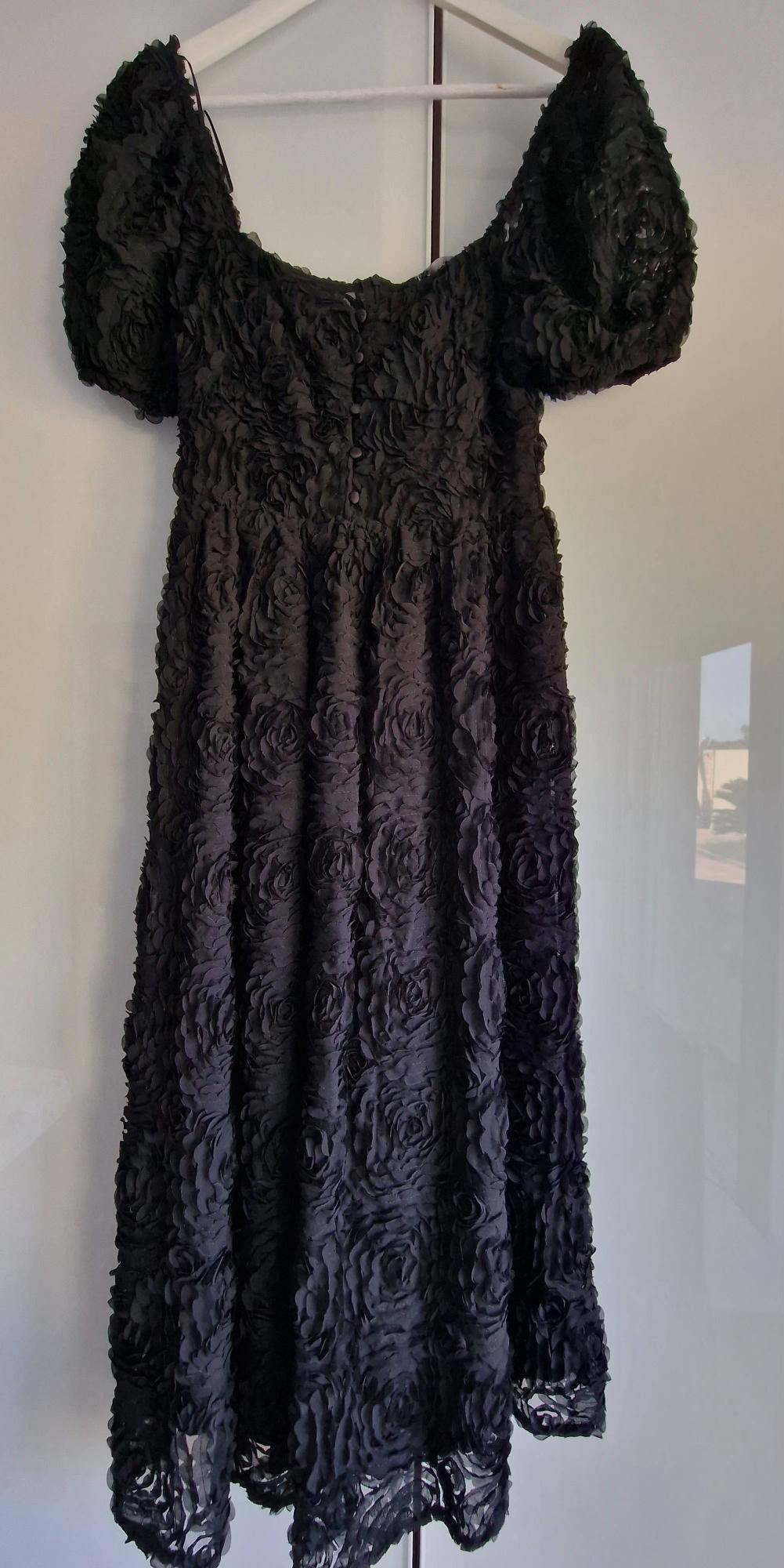 Long black dress covered in embossed roses, featuring short puff sleeves and front buttons, hanging on a white hanger.