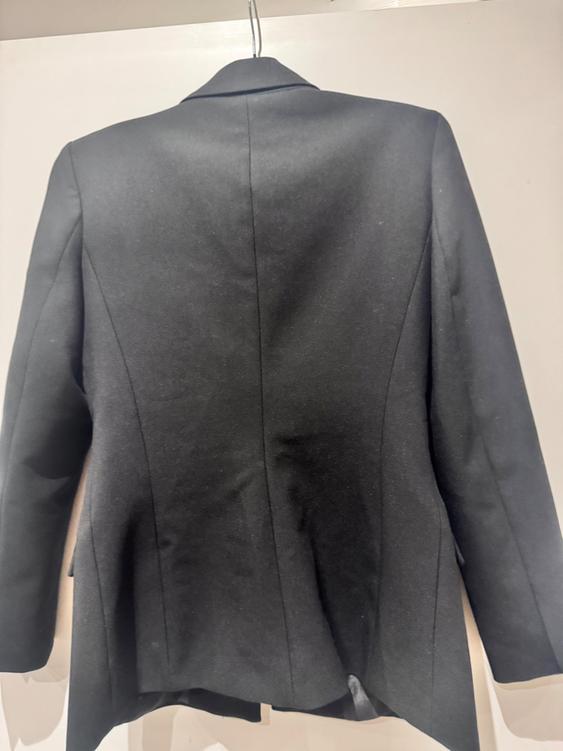 Black jacket hanging on a hanger, viewed from behind, with a white background. The fabric has a smooth and elegant finish.