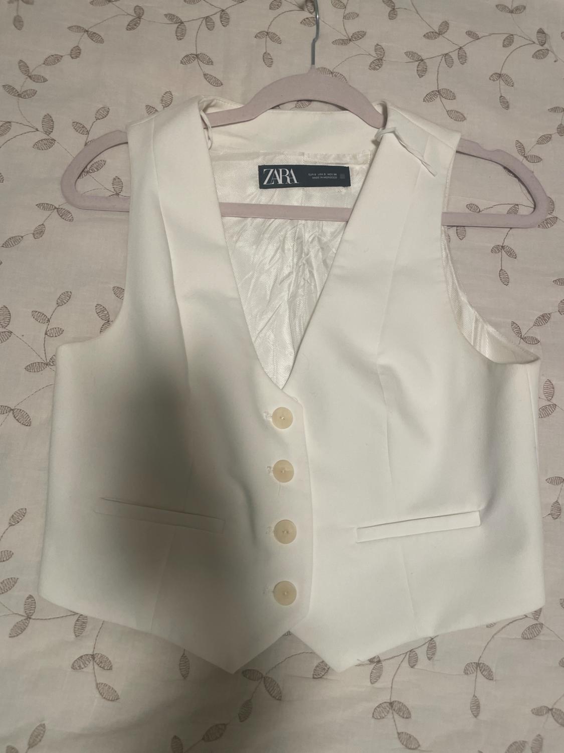 White dress vest with cream buttons, hanging on a hanger over a leaf-patterned fabric. "ZARA" tag visible.