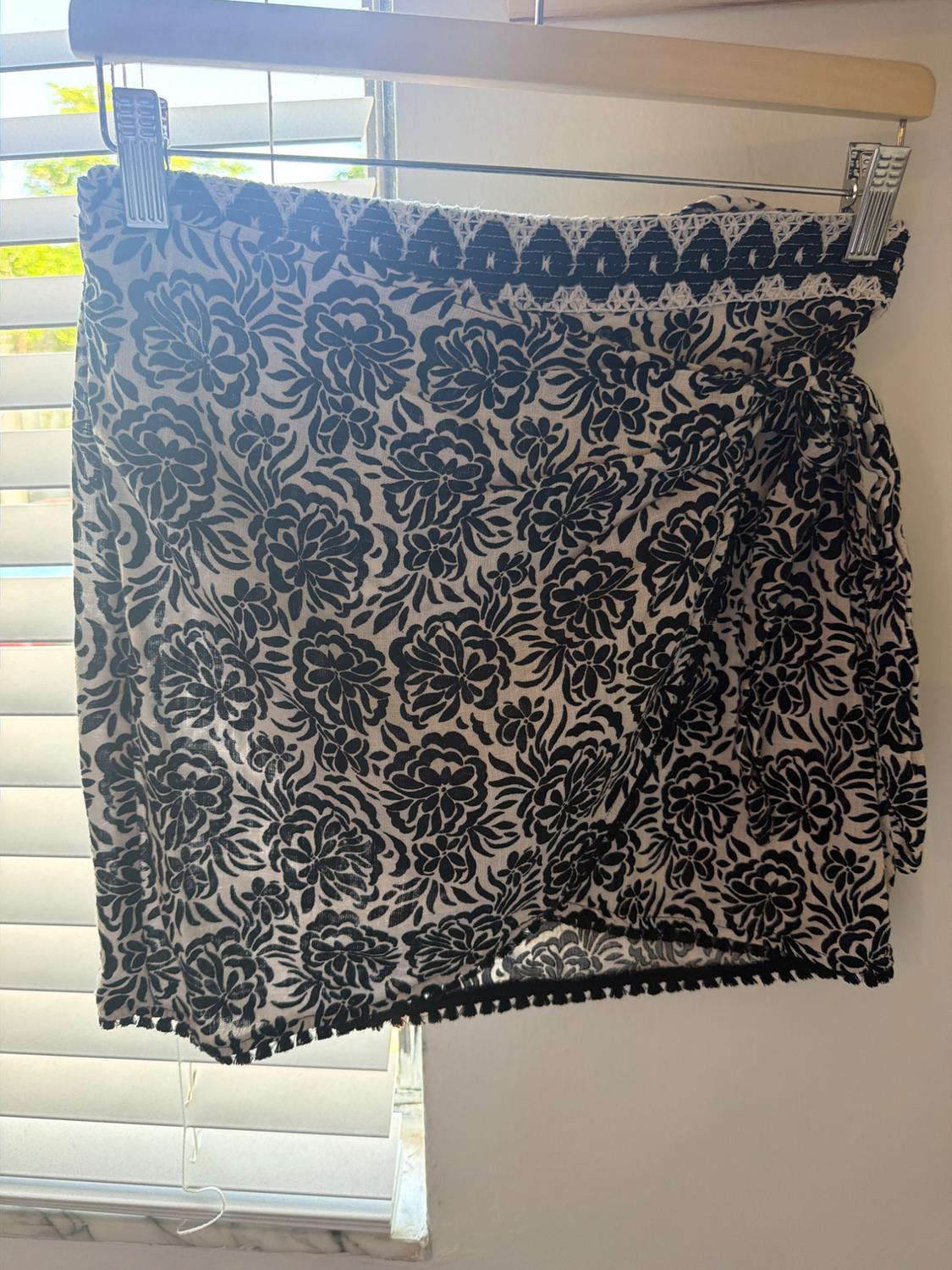 Black and white floral print wrap mini skirt with a side tie and fringe hem, hanging on a hanger in front of a blind.